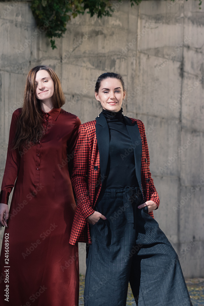 Street style Fashion portrait two charming girls posing. Brown dress, Black and tile red jacket. Summer Spring Fall collection beautiful look. Togetherness, friendship, party concept