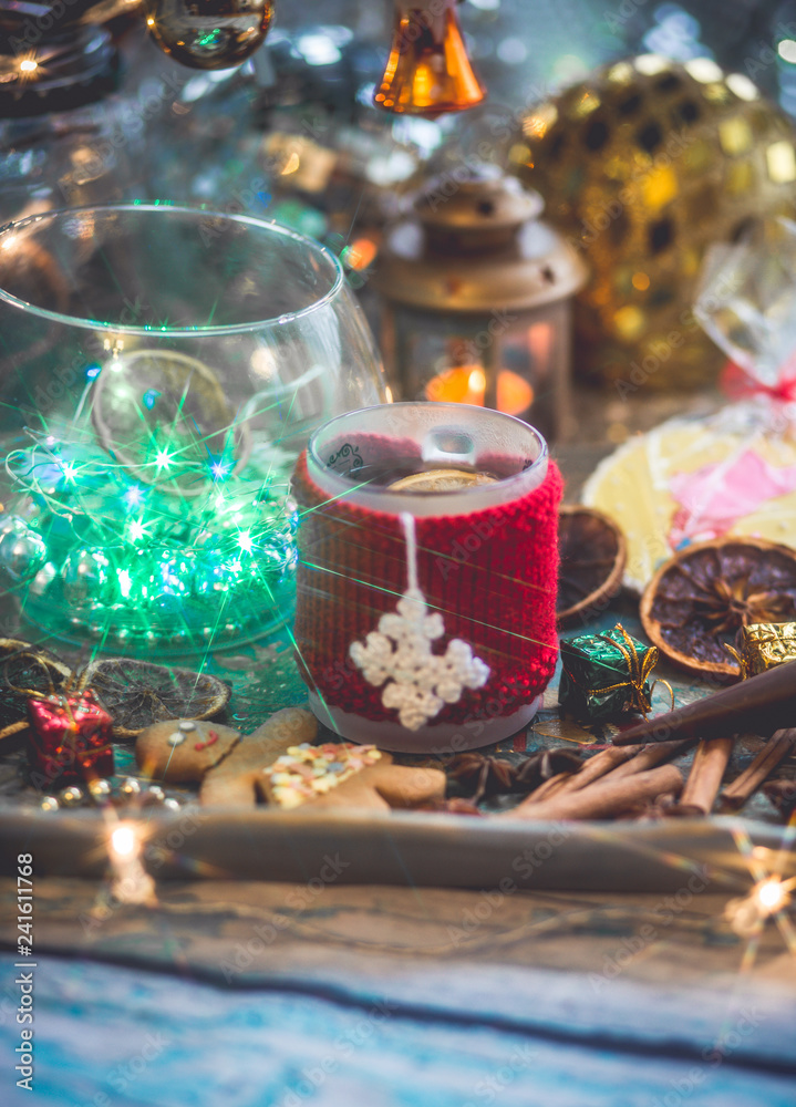 New Year's still life with mug of tea with knitted cup cover, cookies, chocolate, dried fruit and seasonings with lights and bokeh. Photo toned and with vignette