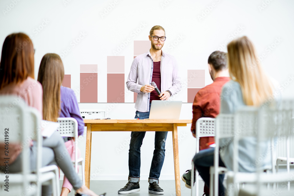 Young male speaker reporting to the audience during the meeting in the conference room with charts on the background