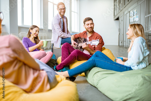 Group of a young coworkers having fun sitting on the colorful poufs playing guitar during the coffee break in the office