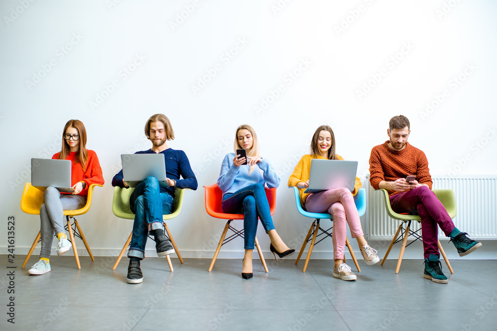 Naklejka premium Young coworkers working with gadgets sitting in a row on the colorful chairs on the white wall background indoors