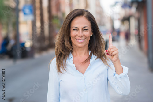 Beautiful middle age hispanic woman at the city street on a sunny day surprised with an idea or question pointing finger with happy face, number one