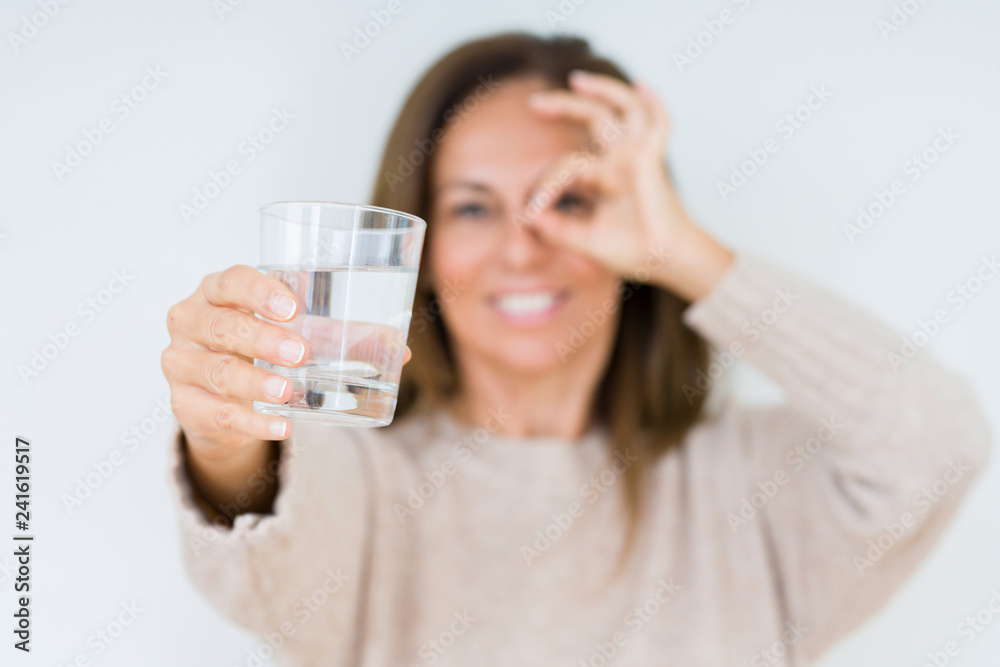 Middle age woman drinking glass of water isolated background with happy face smiling doing ok sign with hand on eye looking through fingers Middle age woman drinking glass of water isolated background with happy face smiling doing ok sign with hand on eye looking through fingers