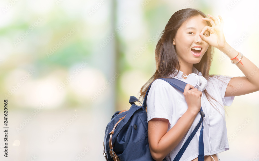 Young asian woman wearing backpack and headphones over isolated background with happy face smiling doing ok sign with hand on eye looking through fingers Young asian woman wearing backpack and headphones over isolated background with happy face smiling doing ok sign with hand on eye looking through fingers