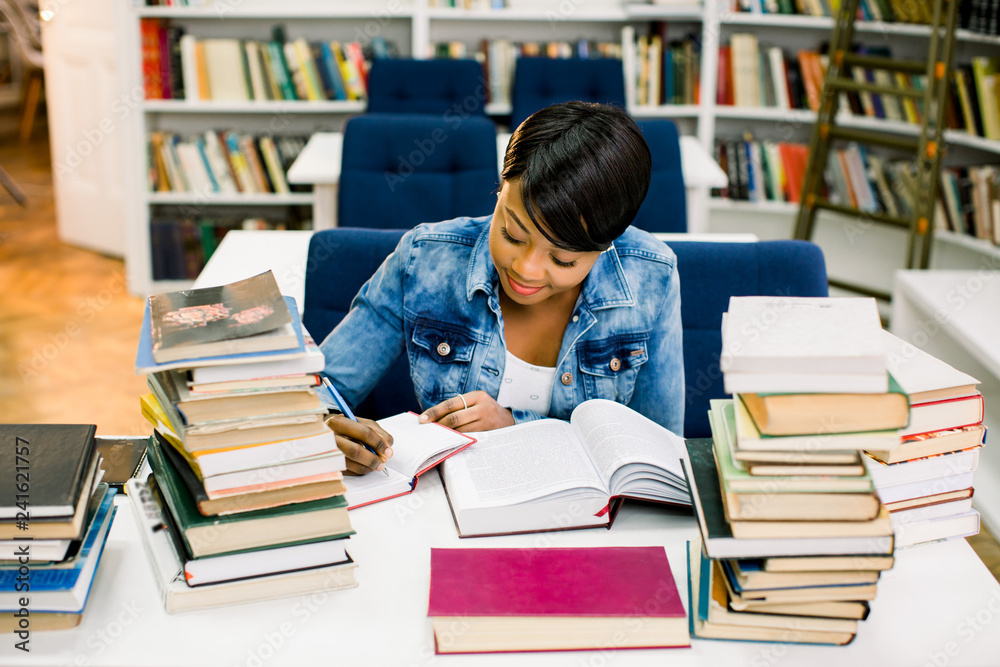 Smart african american young woman dressed in casual wear preparing for studying seminar in library.Intelligent dark skinned hipster student writing and reading Smart african american young woman dressed in casual wear preparing for studying seminar in library.Intelligent dark skinned hipster student writing and reading