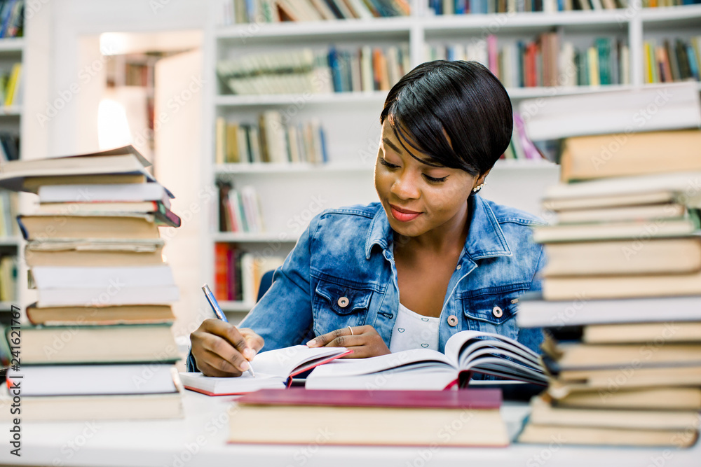 Smart african american young woman dressed in casual wear preparing for studying seminar in library.Intelligent dark skinned hipster student writing and reading
