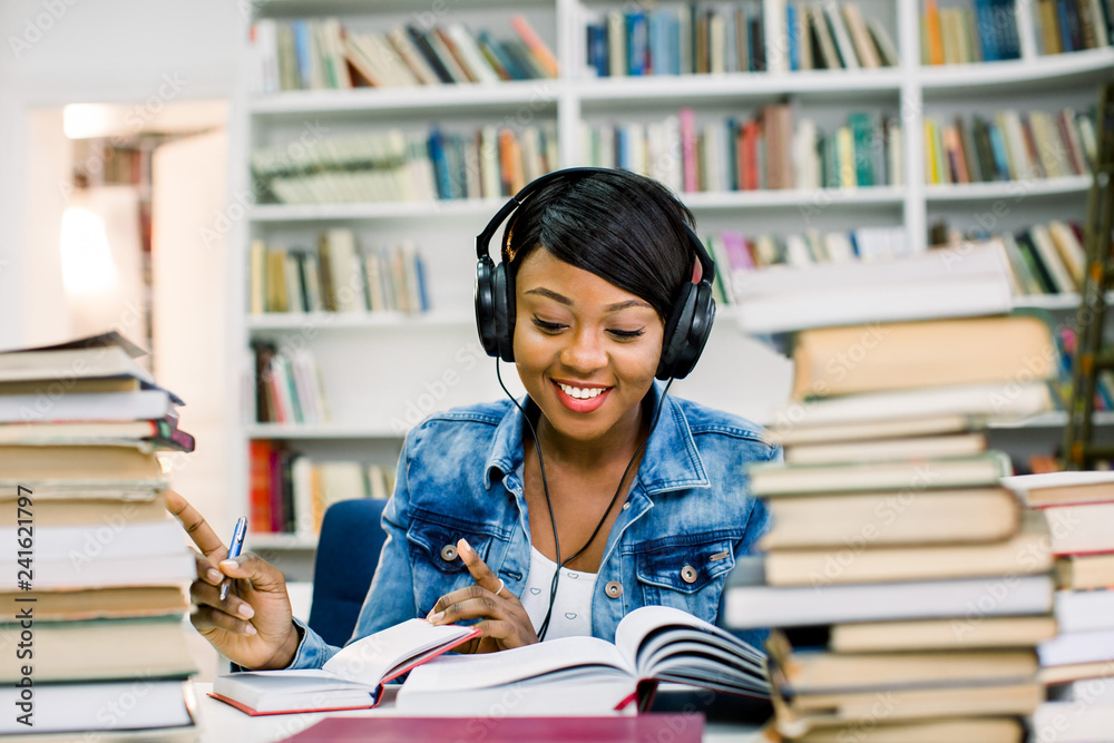Young african smiling woman in a good mood listening to music while studying in a library. Education concept Young african smiling woman in a good mood listening to music while studying in a library. Education concept