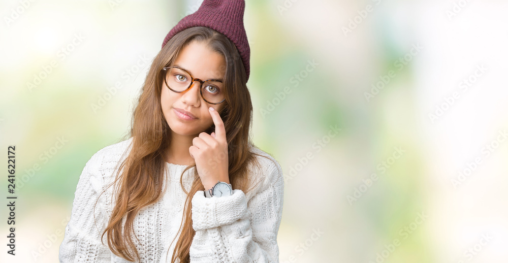Young beautiful brunette hipster woman wearing glasses and winter hat over isolated background Pointing to the eye watching you gesture, suspicious expression Young beautiful brunette hipster woman wearing glasses and winter hat over isolated background Pointing to the eye watching you gesture, suspicious expression