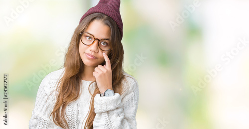 Young beautiful brunette hipster woman wearing glasses and winter hat over isolated background Pointing to the eye watching you gesture, suspicious expression