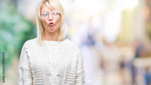 Young beautiful blonde woman wearing winter sweater and glasses over isolated background making fish face with lips, crazy and comical gesture. Funny expression.