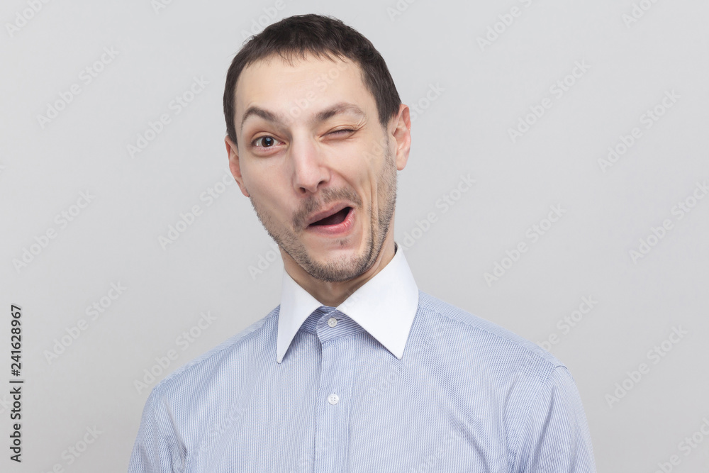Portrait of funny handsome bristle businessman in classic light blue shirt standing, winking and looking at camera with funny face. indoor studio shot, isolated on grey background copyspace. Portrait of funny handsome bristle businessman in classic light blue shirt standing, winking and looking at camera with funny face. indoor studio shot, isolated on grey background copyspace.