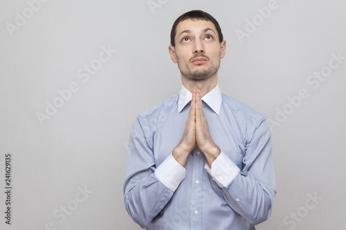 Portrait of praying hopeful handsome bristle businessman in classic blue shirt standing with palm hands and looking at top and pleading God. indoor studio shot, isolated on grey background copyspace.