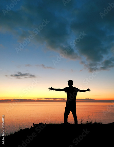 Jeune homme les bras   cart  s sur une falaise face au coucher de soleil sur la mer