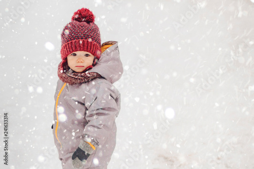 Portrait of a cute baby dressed in a gray jacket and a red hat that walks through the snow covered park enjoying first snow blowing