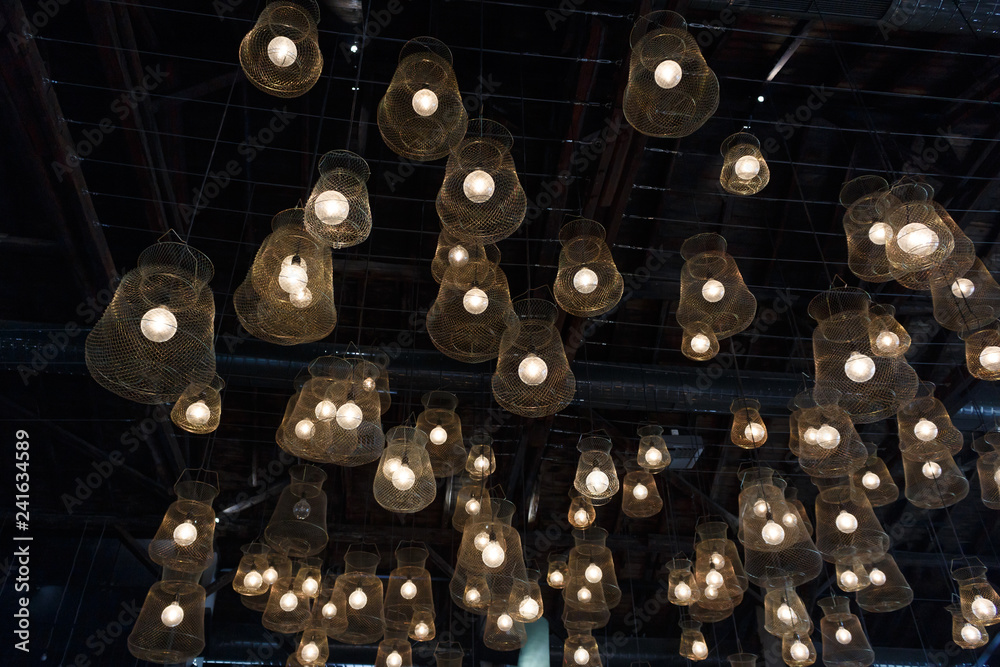 Obraz premium Many vintage incandescent glowing light bulbs with net lampshade hanging from the ceiling on the dark background texture, restaurant interior
