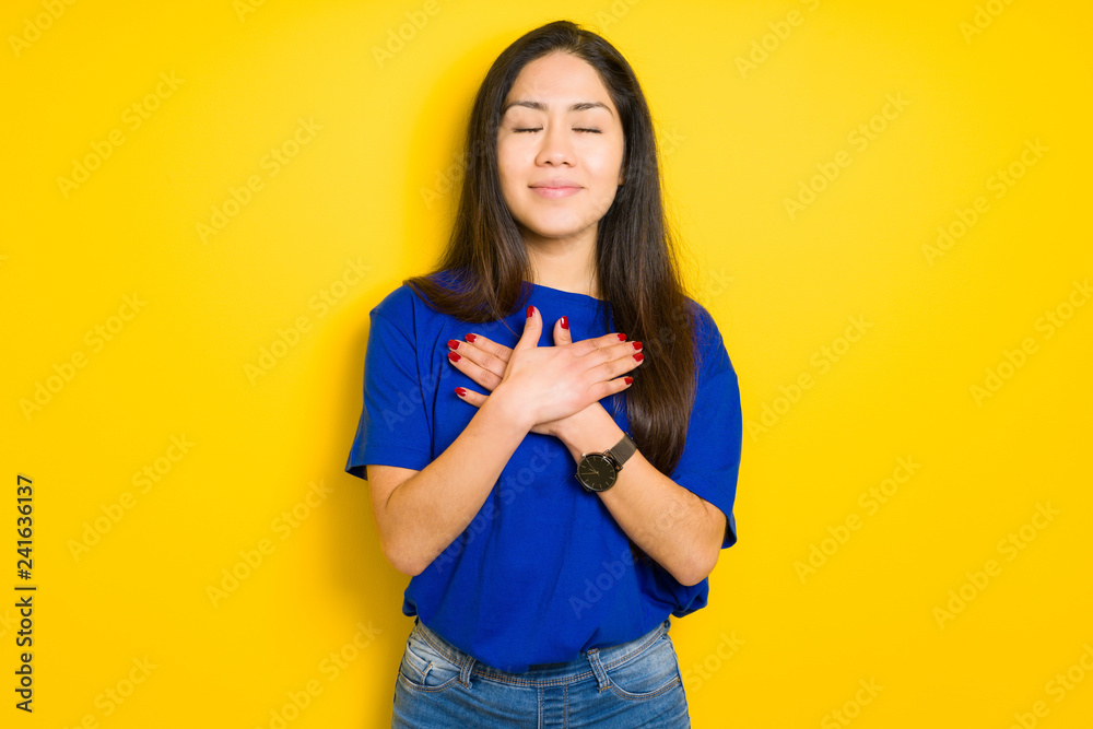 Beautiful brunette woman wearing blue t-shirt over yellow isolated background smiling with hands on chest with closed eyes and grateful gesture on face. Health concept. Beautiful brunette woman wearing blue t-shirt over yellow isolated background smiling with hands on chest with closed eyes and grateful gesture on face. Health concept.