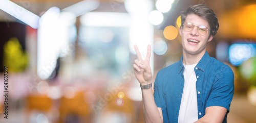 Young handsome man wearing glasses over isolated background smiling with happy face winking at the camera doing victory sign. Number two.