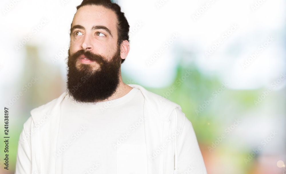Young man with long hair and beard wearing sporty sweatshirt smiling looking side and staring away thinking. Young man with long hair and beard wearing sporty sweatshirt smiling looking side and staring away thinking.