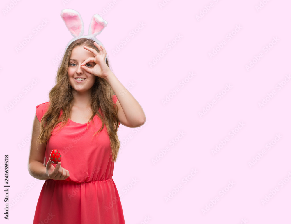 Young blonde woman wearing easter bunny ears with happy face smiling doing ok sign with hand on eye looking through fingers Young blonde woman wearing easter bunny ears with happy face smiling doing ok sign with hand on eye looking through fingers