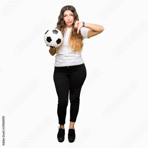 Young adult woman holding soccer football ball with angry face, negative sign showing dislike with thumbs down, rejection concept