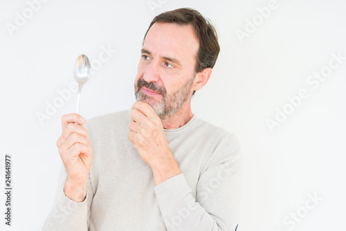 Senior man holding silver spoon over isolated background serious face thinking about question, very confused idea