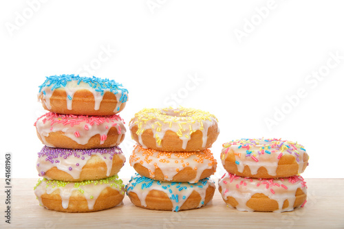 Many colorful frosted cake donuts with candy sprinkles stacked in rows laying on wood table isolated on white background. Fun festive party food.