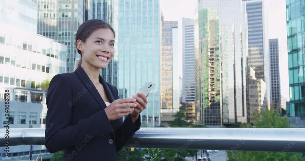 Smiling young businesswoman holding mobile phone while looking away. Confident female professional is standing by modern buildings. She is wearing suit in city. Smiling young businesswoman holding mobile phone while looking away. Confident female professional is standing by modern buildings. She is wearing suit in city.