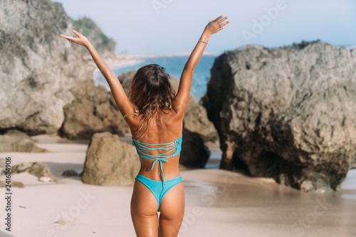 Backside view of slender girl in swimsuit with raised arms up stands on sandy beach with rocks