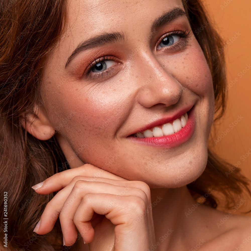 Cute caucasian brunette woman with curly hairstyle on orange background. Fresh clean skin with evening make up and freckles. Hands near her face. Close up portrait Cute caucasian brunette woman with curly hairstyle on orange background. Fresh clean skin with evening make up and freckles. Hands near her face. Close up portrait