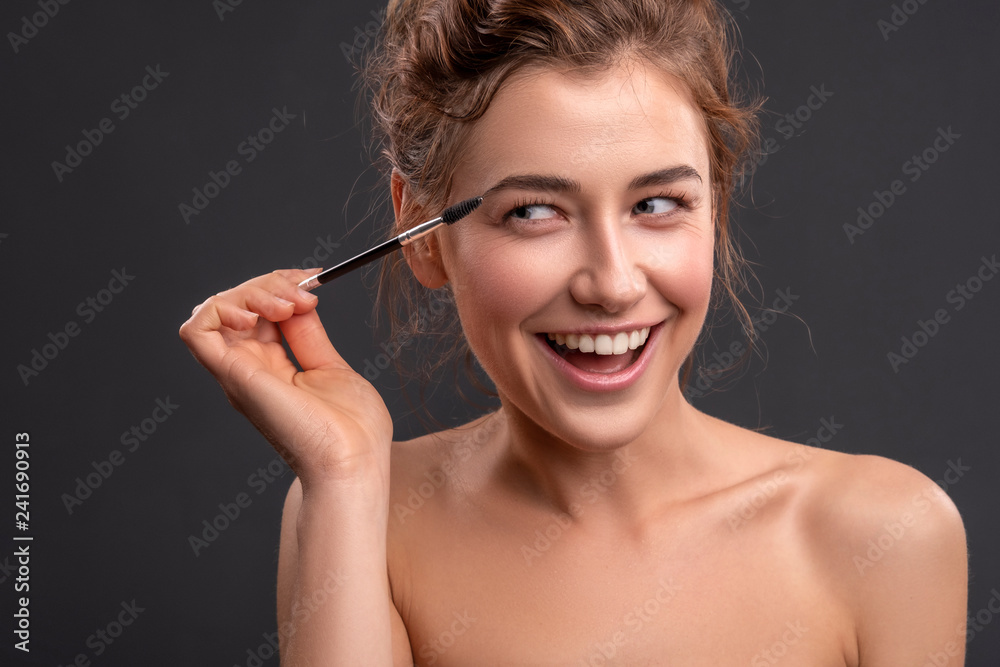 Obraz premium Beautiful brunette caucasian young woman prepare herself, applying mascara on her eyes with a brush. Clean, fresh, natural, flawless skin. Soft smile on her face. Close up on a neutral background