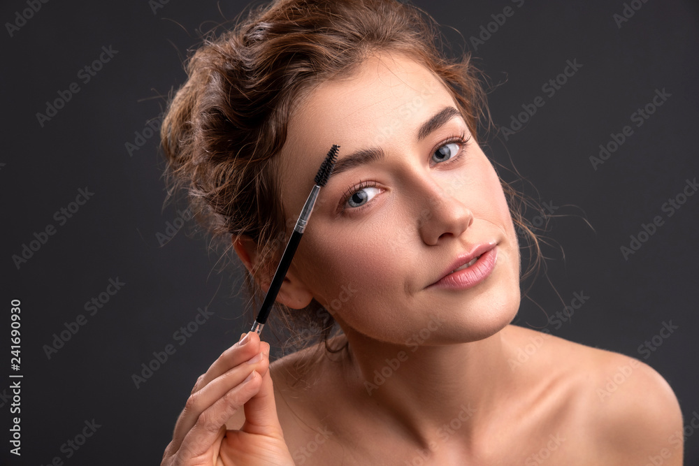 Fototapeta premium Beautiful brunette caucasian young woman prepare herself, applying mascara on her eyes with a brush. Clean, fresh, natural, flawless skin. Soft smile on her face. Close up on a neutral background