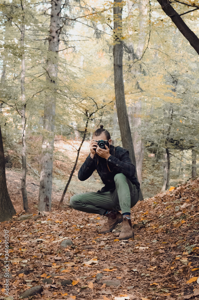 Hipster Travel Lifestyle concept: stylish bearded man, dressed in a shirt and with a hairdo Top Knot with retro photo camera in hands takes pictures in the woods