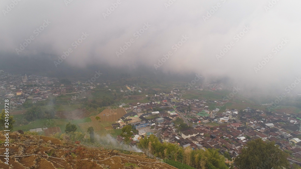 asian town in mountains among agricultural land, rice terraces, fog and clouds Java Indonesia. mountains with farmlands, rice fields, village, fields with crops, trees. Aerial view farm lands on