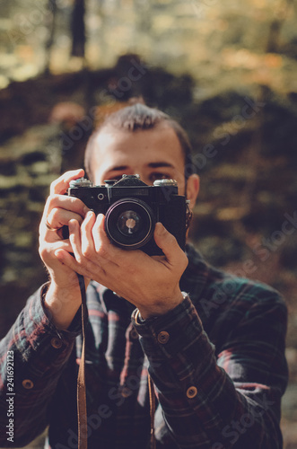 Nature Photography Concepts Professional photographer: stylish bearded man, dressed in a shirt and with a hairdo Top Knot with retro photo camera in hands takes pictures in the woods