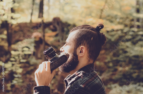 Hipster Travel Lifestyle concept: stylish bearded man, dressed in a shirt and with a hairdo Top Knot with retro photo camera in hands takes pictures in the woods