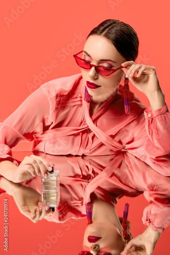 glamorous woman in blouse and red sunglasses posing with perfume bottle and mirror reflection isolated on living coral
