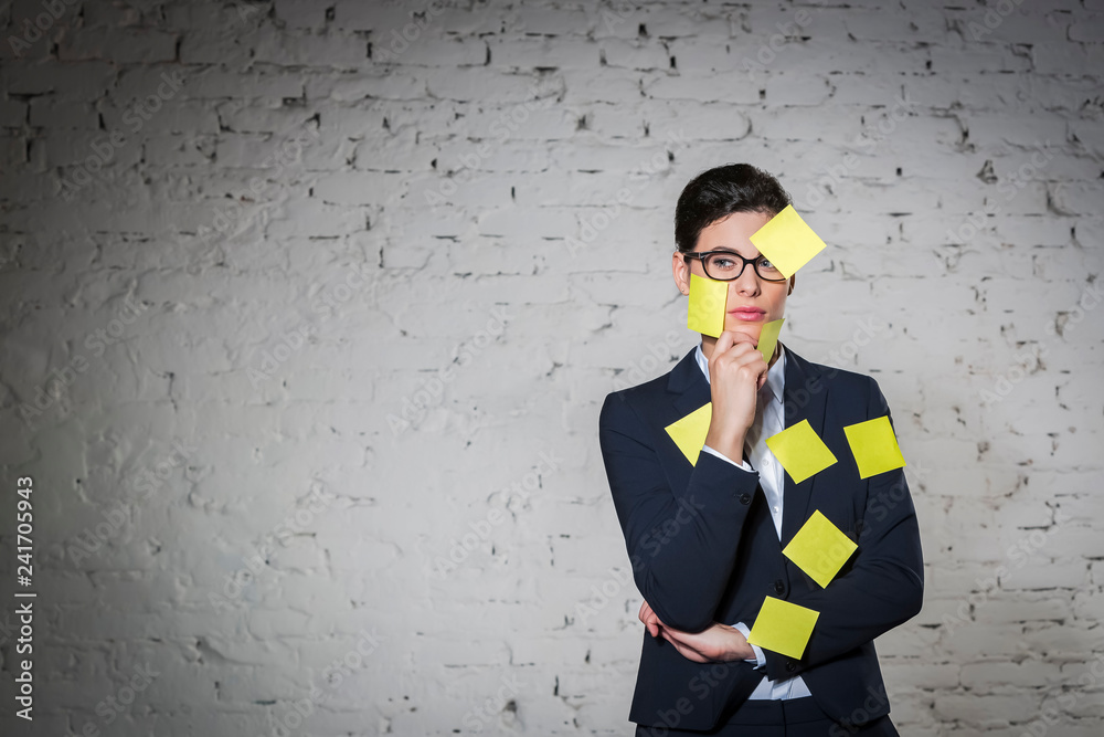 Thoughtful businesswoman with blank yellow adhesive notes on face and suit against wall at office Thoughtful businesswoman with blank yellow adhesive notes on face and suit against wall at office