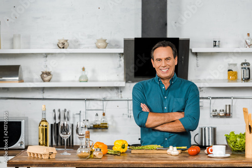 smiling handsome middle aged man standing with crossed arms and looking at camera in kitchen