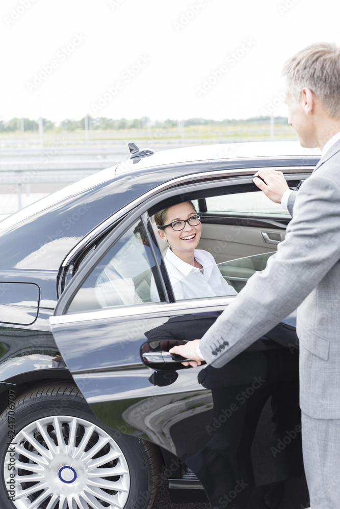 Executive holding car door for smiling colleague against sky Executive holding car door for smiling colleague against sky