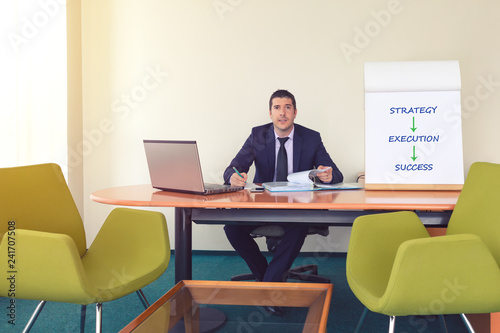 Confident young team leader or manager sitting at desk next to laptop and flip chart smiling while working with documents and taking notes – concept of business strategy, success – warm filter © DanRentea