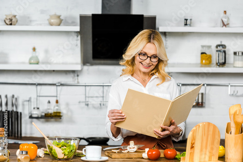 smiling attractive mature woman reading recipe book for cooking in kitchen