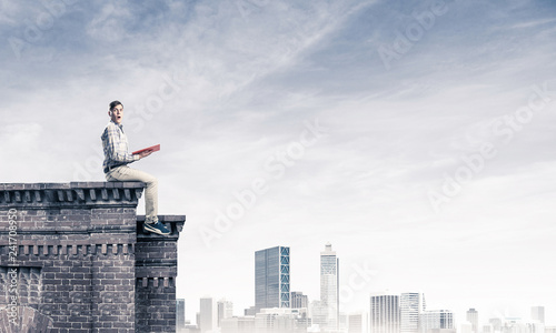 Man on roof edge reading book and cityscape at background © adam121