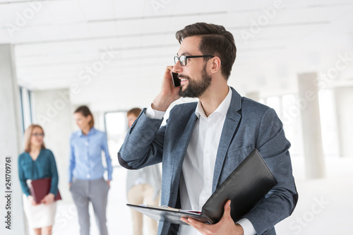 Businessman with file talking on mobile phone against colleagues in new office