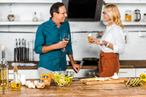 selective focus of mature wife and husband holding glasses of wine and looking at each other in kitchen