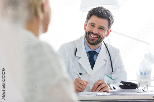 Obraz na płótnie Smiling doctor discussing with mature patient while writing prescription at desk