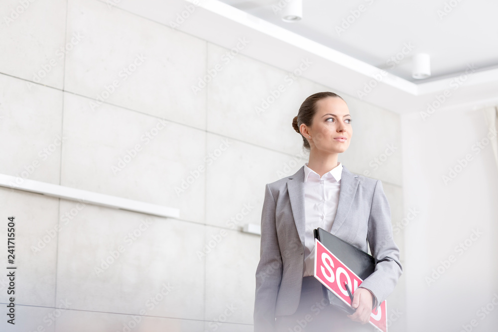 Confident young saleswoman looking away while standing with sold placard and document against wall in apartment Confident young saleswoman looking away while standing with sold placard and document against wall in apartment
