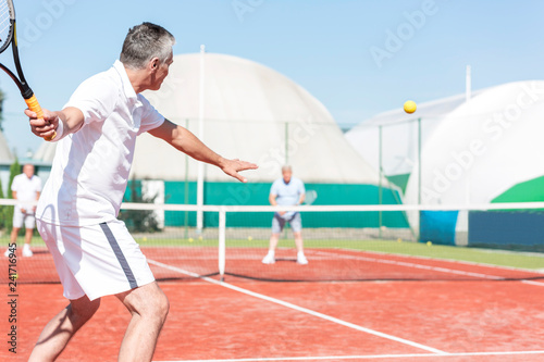 Man swinging tennis racket while playing doubles match on red court during summer weekend