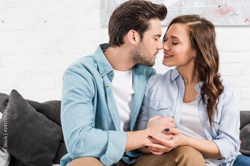 beautiful couple sitting on couch and holding hands at home while woman looking at camera beautiful couple sitting on couch and holding hands at home while woman looking at camera