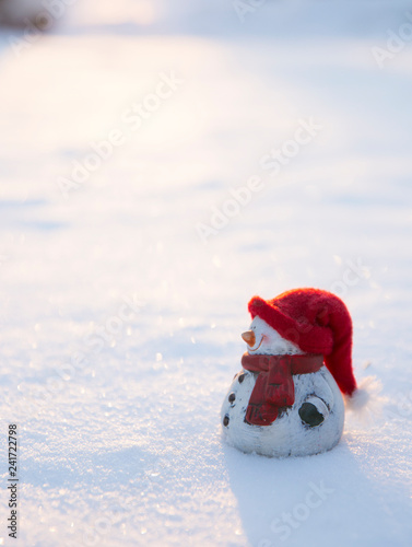 White snowman figure with warm hat and scarf standing in snow, sun setting outdoors in winter, lot of copy space. © FotoHelin