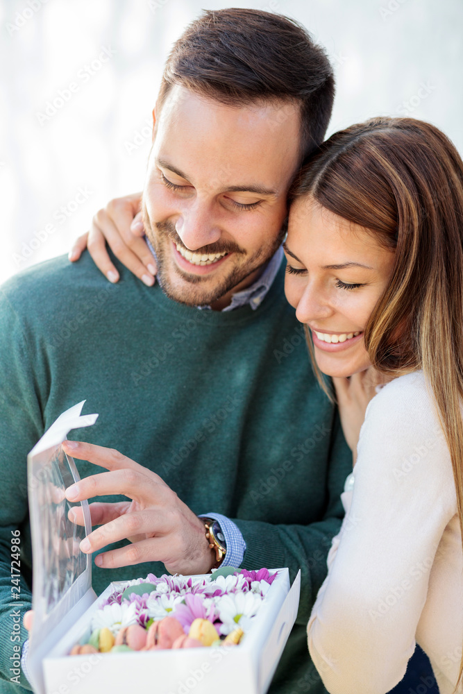 Fototapeta premium Happy young couple celebrating Valentine's Day. Woman is hugging her boyfriend or husband as he is opening a gift box